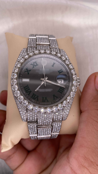 Angle view of iced out moissanite watch with diamond bezel, silver strap, black dial, green Roman numerals, and date display.